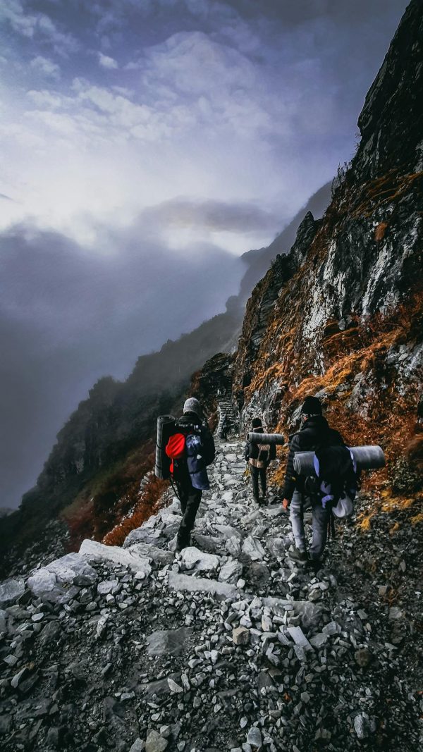 Back view of unrecognizable travelers with backpacks climbing up mountain slope on cloudy day