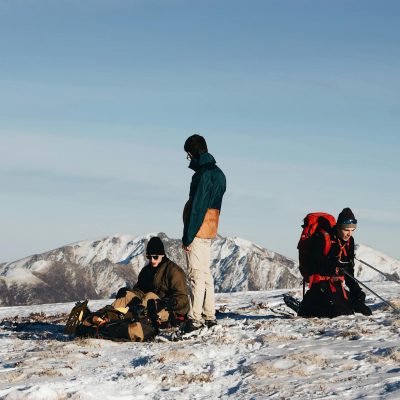 Group of travelers in outerwear with backpacks and hiking equipment resting on snowy ground during expedition against cloudless blue sky and mountain peaks
