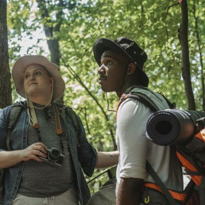 Plus size guy with photo camera and black young bearded man with backpack and tourist mat standing near tent and looking up